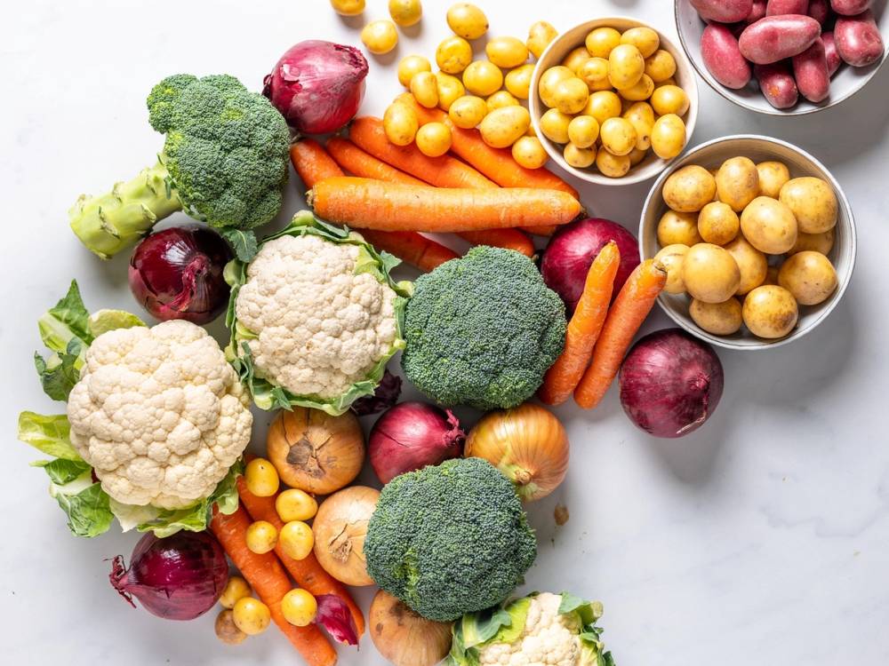 Et utvalg av ulike ferske grønnsaker som brokkoli, gulrøtter, rødløk, blomkål og kikerter, spredt ut på en hvit bakgrunn.