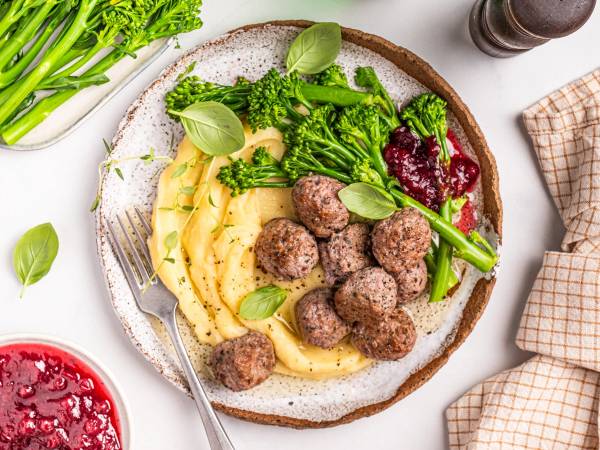 Kjøttboller med potetmos og grønnsaker på en tallerken, servert med grønne urter og en liten skål med rød saus.