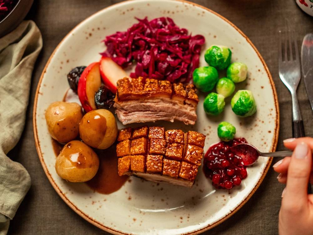 En tallerken med svinekjøtt, kokte poteter, rødkål, grønne erter og tyttebær, med en person som holder en skje med tyttebær på høyre side.