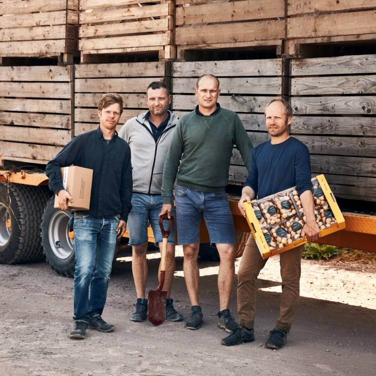 Fire personer står sammen foran en trevegg, hvorav en holder en eske og en annen et brett med bilder eller kort.