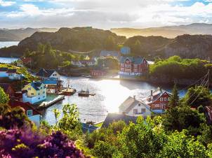 En liten havneby med fargerike hus og båter ved vannet, omgitt av grønne åser og fjell i bakgrunnen.