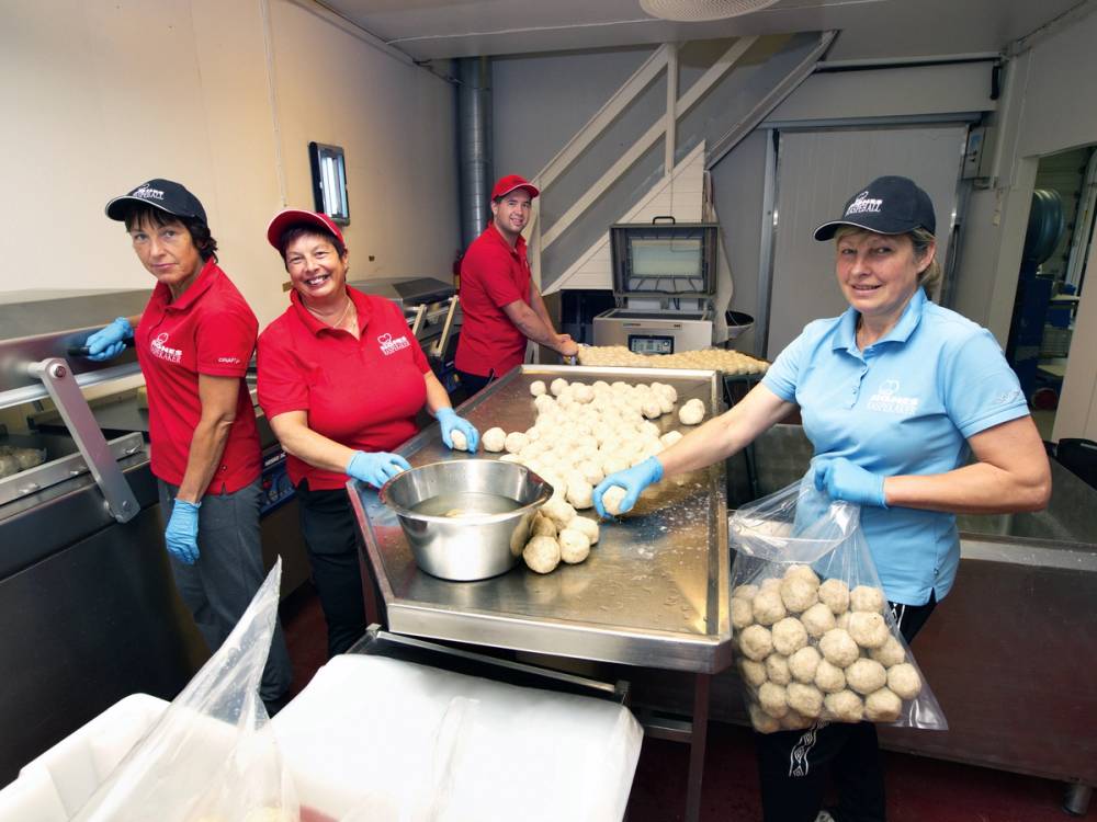 Fire personer i arbeidsdrakt jobber med konservering i et kjøkken, med en maskin og kuler av mat på bordet, i Hordaland.