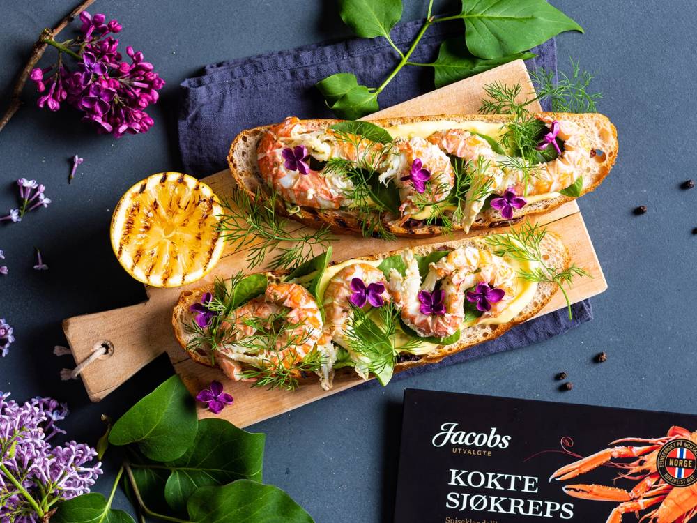 To baguetter med sjøkreps, urter og spiselige blomster på en skjærebrett, omgitt av blomster og grønt blader.