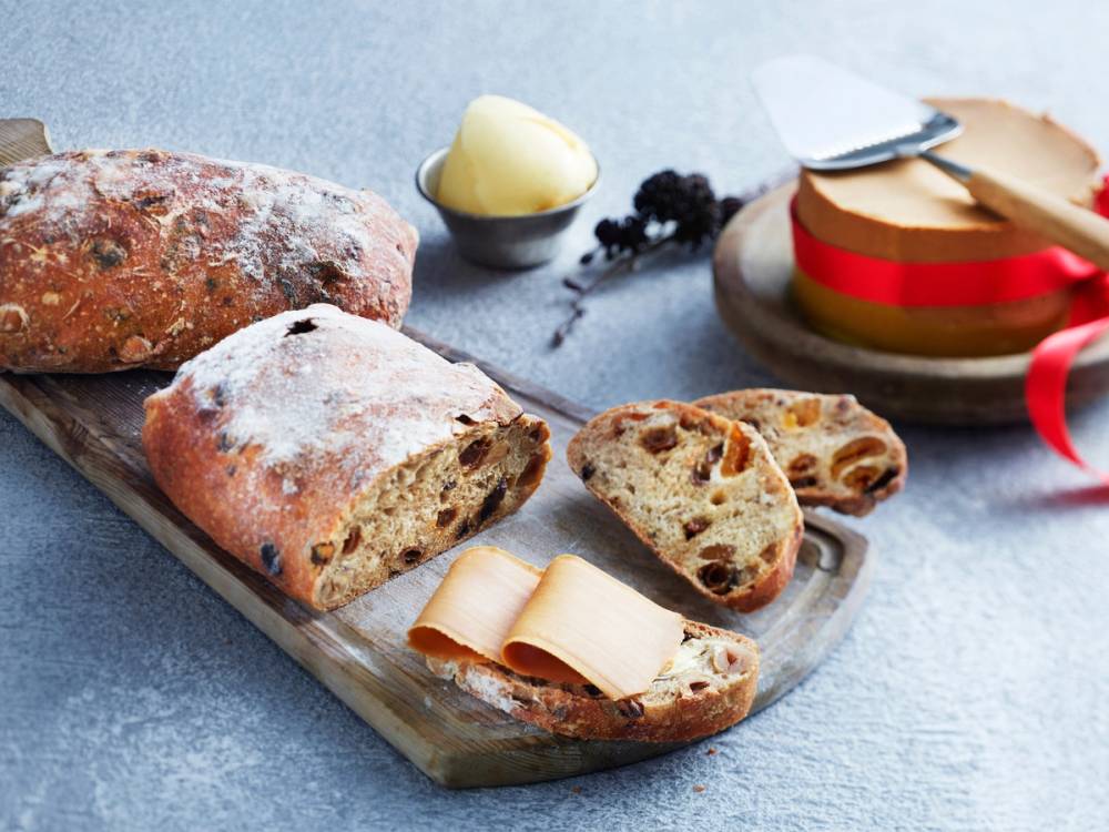 Frukt-nottebrød med brunost på et skjærebrett, omgitt av ostebiter og en liten skål med smør eller smøraktig pålegg.