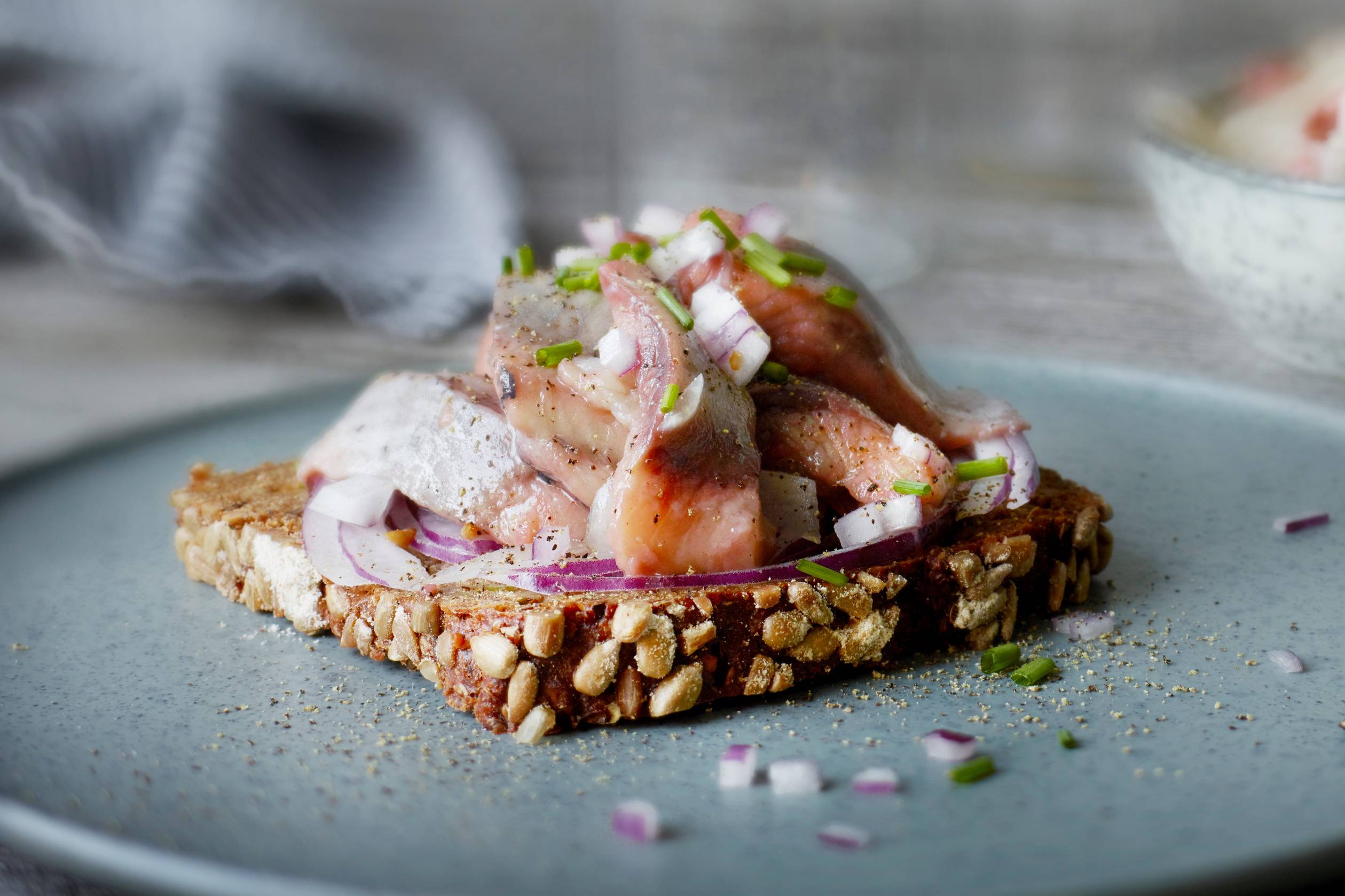 En skive med hjemmelaget pålegg med kylling, løk og grønne urter, ser ut til å være på en tallerken med nøtter og frø på, på et kjøkkenbord.