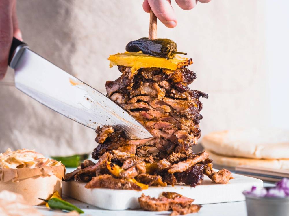 En person skjærer i en stor, høy shawarma med kjøtt og grønnsaker på en skjærefjøl.