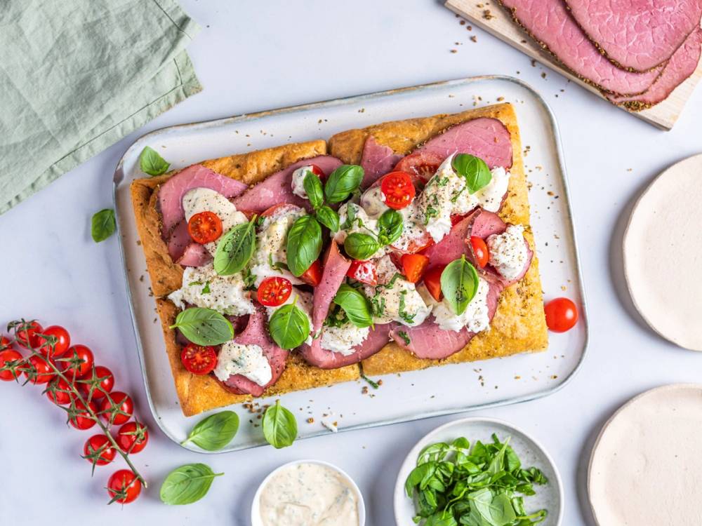 Pastrami-sandwich med grønne blader, cherrytomater og hvitløkssaus på en hvit tallerken, omgitt av tomater og urter.