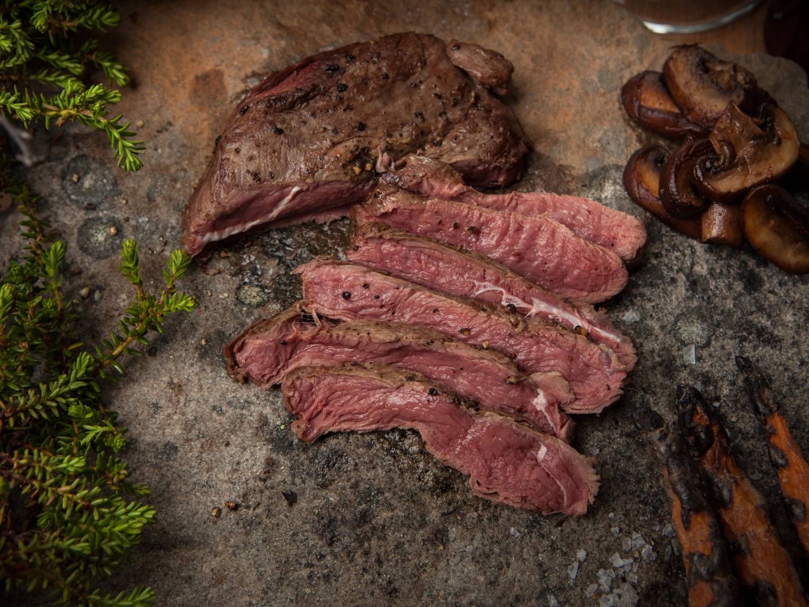 Skiver av viltkjøtt lagt ut på en stein med sopp og urter rundt, trolig til matlaging.