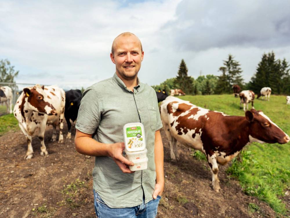 En person står på en gård med kyr, og holder en pakke med melk i hånden, med grønne trær og en åpen himmel i bakgrunnen.