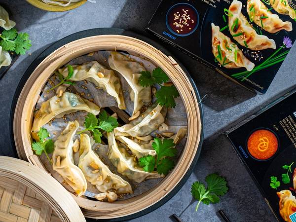 Frosne gyoza-pakker og en bambusdampetallerken med ferdigkokte gyoza, pyntet med koriander, på et mørkt bord.