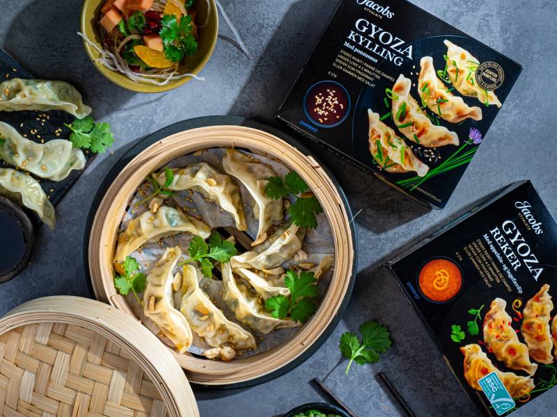 Frosne gyoza-pakker og en bambusdampetallerken med ferdigkokte gyoza, pyntet med koriander, på et mørkt bord.