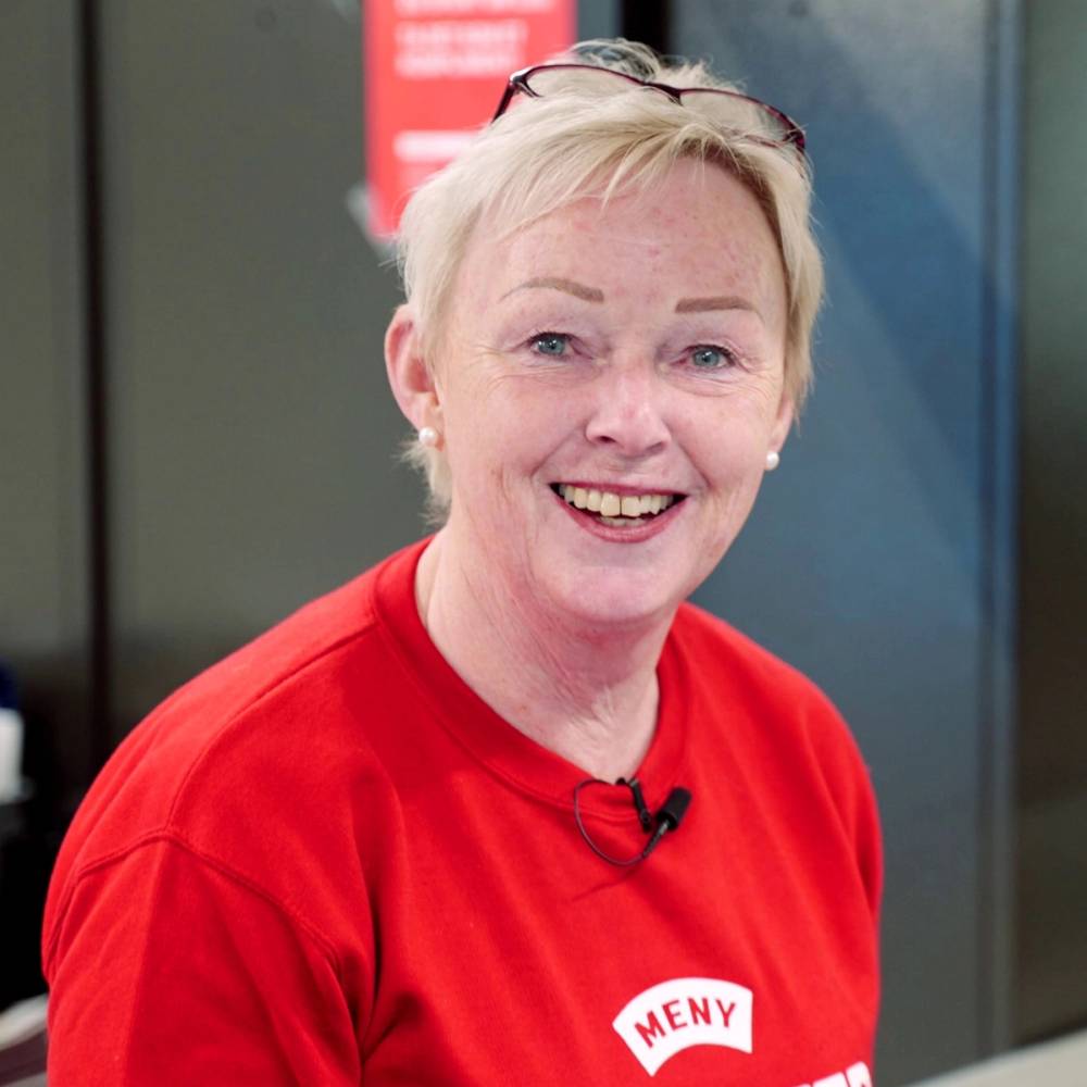 En person med lyst hår som bruker en rød genser med MENY-logo, står foran en blå vegg og en rødt skilt i bakgrunnen.