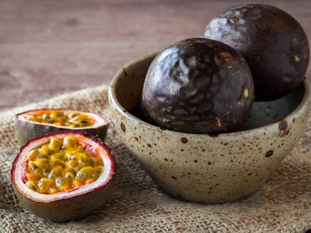 To pasjonsfrukter, en hel og en halvert, i en keramikkbolle på et treverk, med den halverte frukten som viser frø og gult innhold.