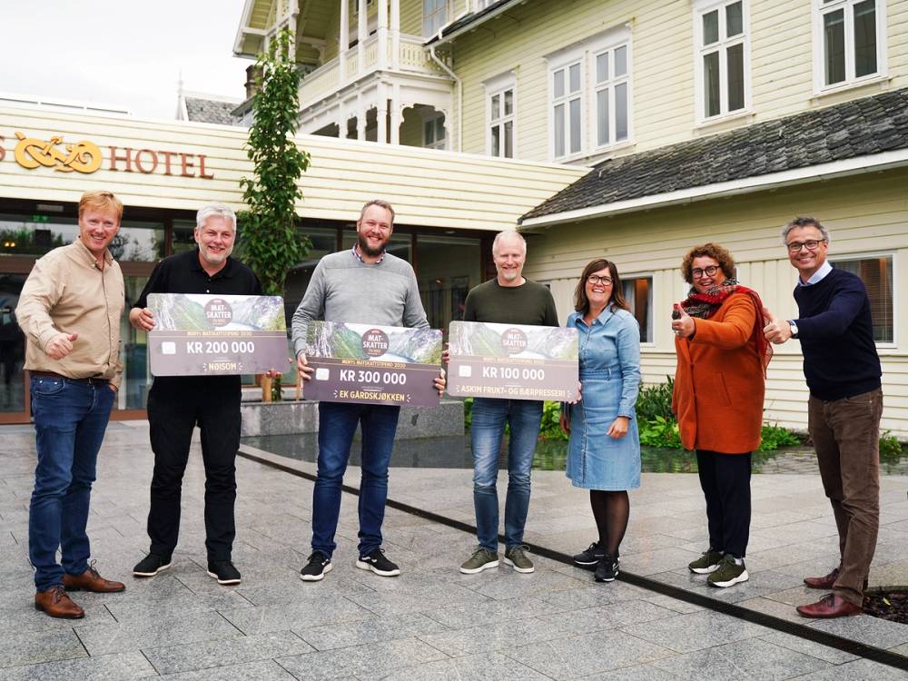 Syv personer står foran et hotell og holder store sjekk- eller diplombilder, trolig for å feire vinnere av Matskattstipendet 2020.