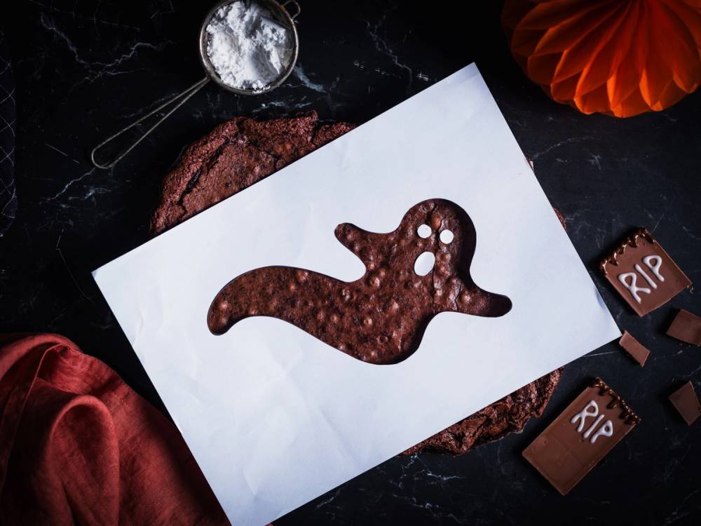 En kake med en sjiraffform laget av sjokolade, plassert på et hvitt ark på en svart overflate med Halloween-dekorasjoner rundt.