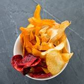 En liten hvit bolle med ulike typer chips og snacks, inkludert potetgull og grønnsakschips, på en mørk overflate.