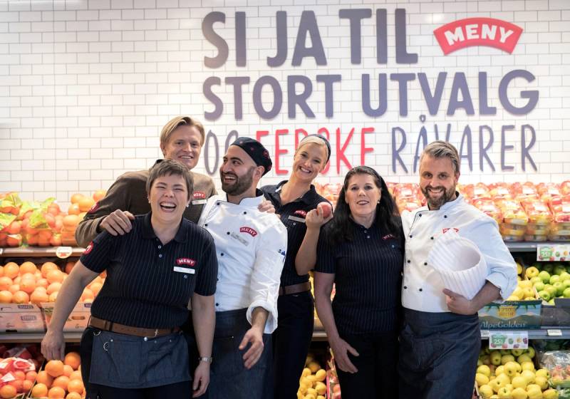 Ansatte foran inngangen til en butikk med en vegg som har tekst om utvalg og råvarer, og frukt i bakgrunnen.