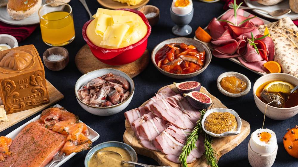 Et julefrokostbord med ulike retter, pålegg, egg, ost, og julepynt, sett på et mørkt bord.