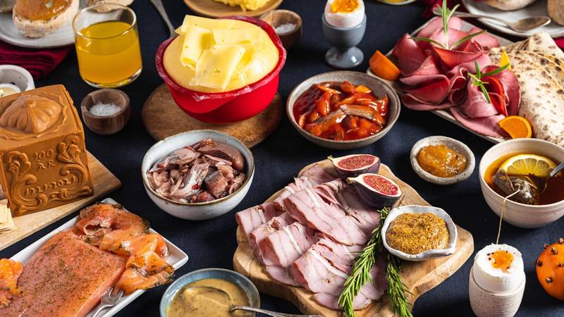 Et julefrokostbord med ulike retter, pålegg, egg, ost, og julepynt, sett på et mørkt bord.