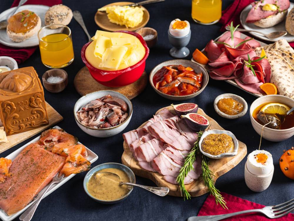 Et julefrokostbord med ulike retter, pålegg, egg, ost, og julepynt, sett på et mørkt bord.