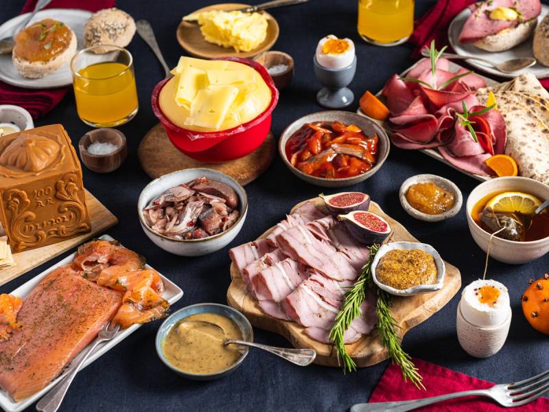 Et julefrokostbord med ulike retter, pålegg, egg, ost, og julepynt, sett på et mørkt bord.
