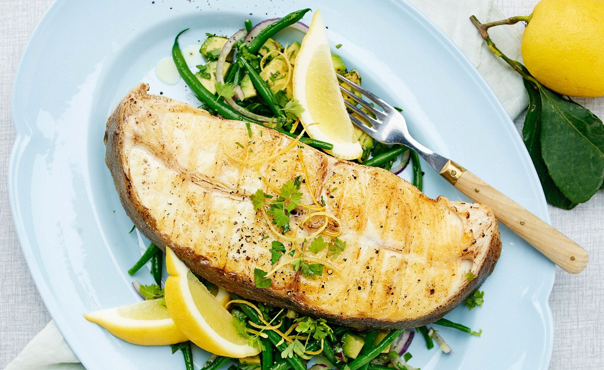 Grillet kveite med aspargesbønner og sitronbåter på en hvit tallerken, med en sitron og en grønn gren i bakgrunnen.