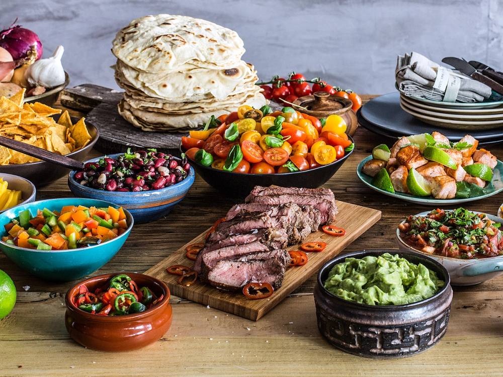 Et utvalg av taco-tilbehør med kjøtt, grønnsaker, salsa og tortillaer på et bord.