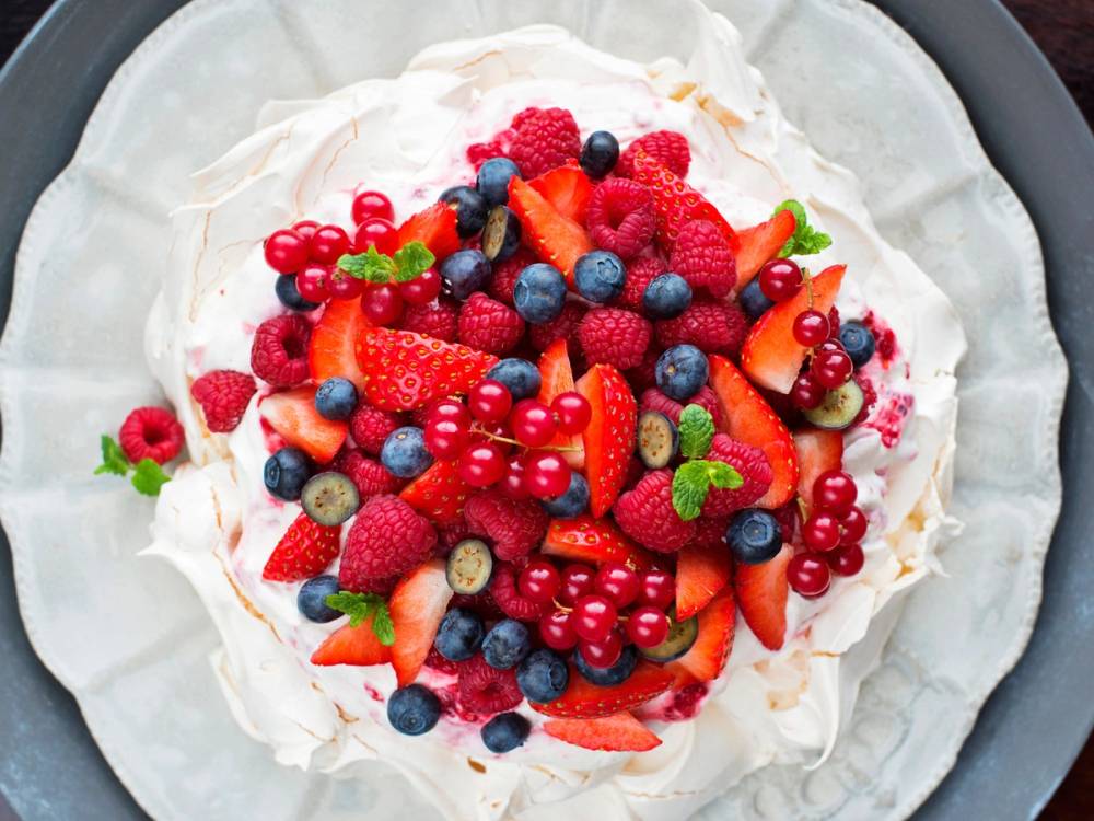 En pavlova dekket med ulike friske bær som jordbær, blåbær, bringebær og rips, på en hvit tallerken.