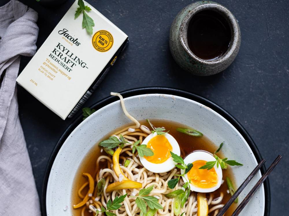 En bolle med nudler, halverte egg og friske urter, med en kopp saus og en pakke Jacobs utvalgte saus og kraft i bakgrunnen.