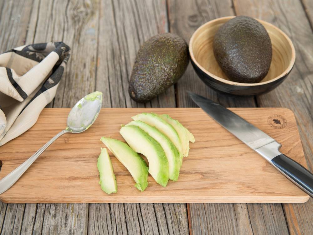Skrellet avocado i skiver på et skjærebrett med en kniv og en skje, og to hele avokadoer i bakgrunnen.