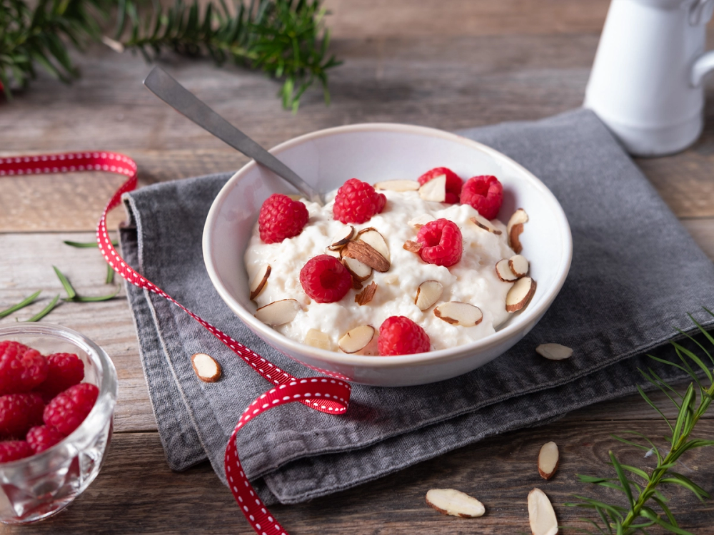 Risgrøt med bringebær og mandelflak i en hvit bolle på et bord med en skje oppi.