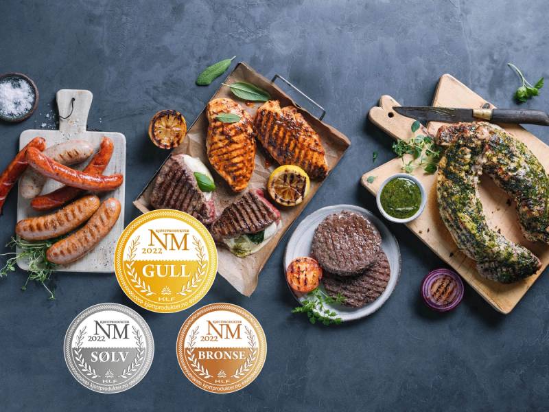 Ulike grillet kjøtt, pølser og fisk på fat og tallerkener med tilbehør, samt medaljer for NM i grilling, på et mørkt bord.