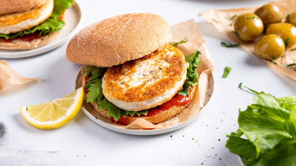 Fiskekaker i et hamburgerbrød med salat og tomat, servert med sitron og grønne oliven på siden.