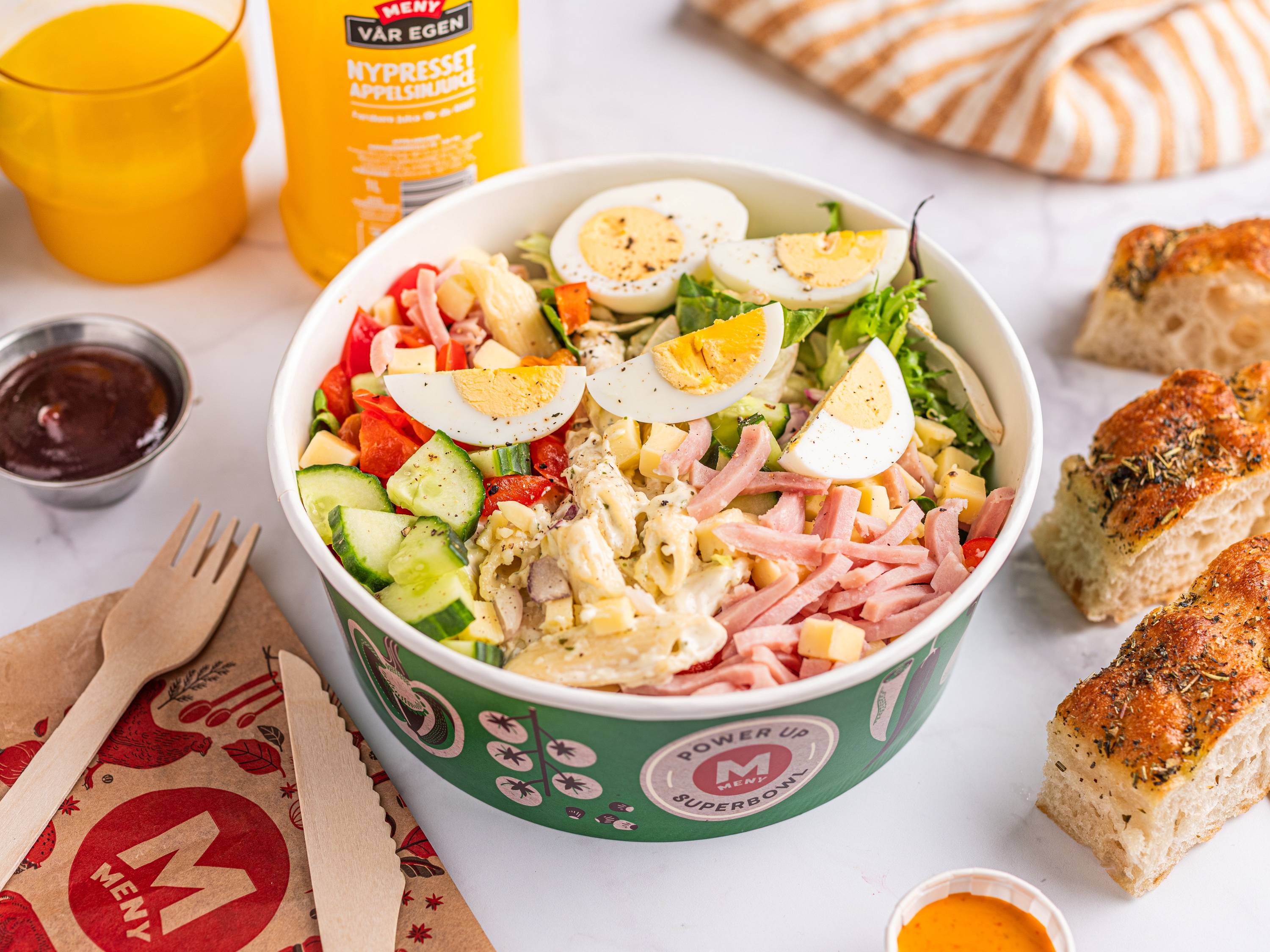 En salat med kokte egg, skinke, agurk, tomat og salat i en grønn og hvit papirskål, omgitt av brød og dressingflasker.