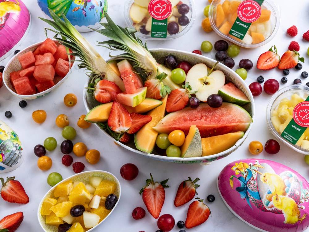 Fargerik påskeegg og frukt på et bord, med vannmelon, jordbær og druer, omgitt av små egg og godteri.