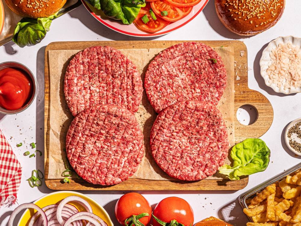 Fire rå hamburgerkjøttdeigformer på en skjærefjøl med grønnsaker og tilbehør rundt omkring.