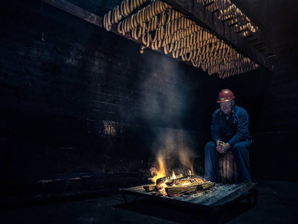 En person i arbeid i et eldre eldhus med åpen ild og tradisjonelle trestrukturer i taket, sannsynligvis i Hordaland.