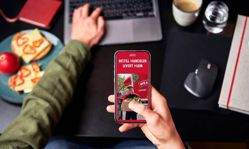 En person holder en mobiltelefon med en meny for hjemlevering av mat, mens de sitter ved et skrivebord med en datamaskin og andre kontorrekvisita.