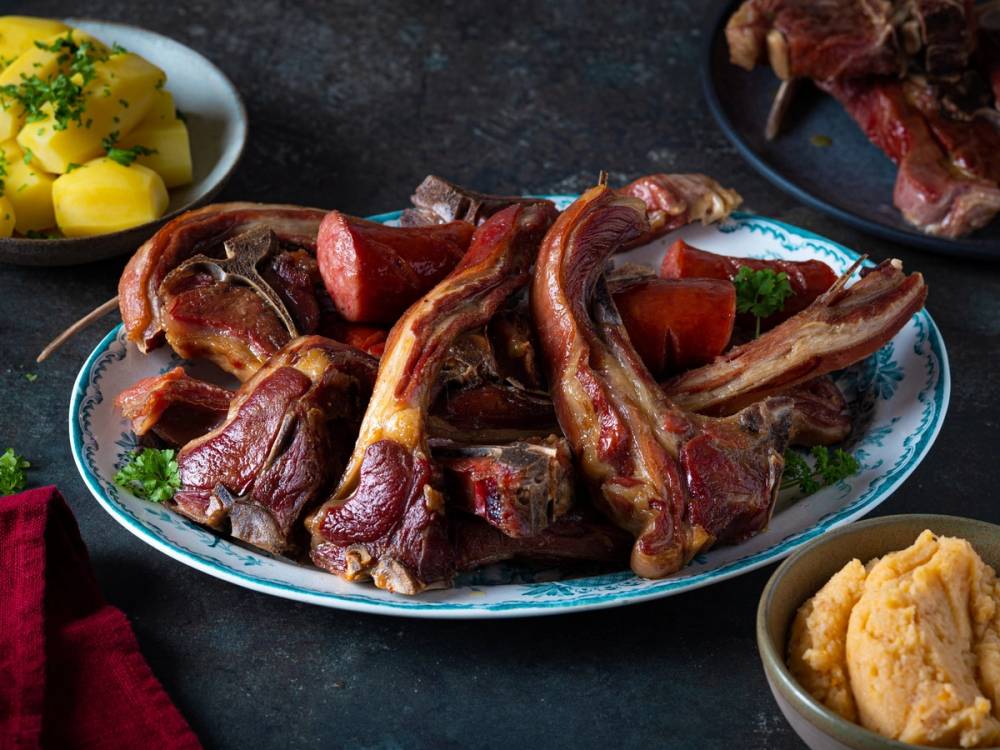 Pinnekjøtt med kålrotstappe på et serveringsfat, omgitt av grønnsaker og tilbehør.