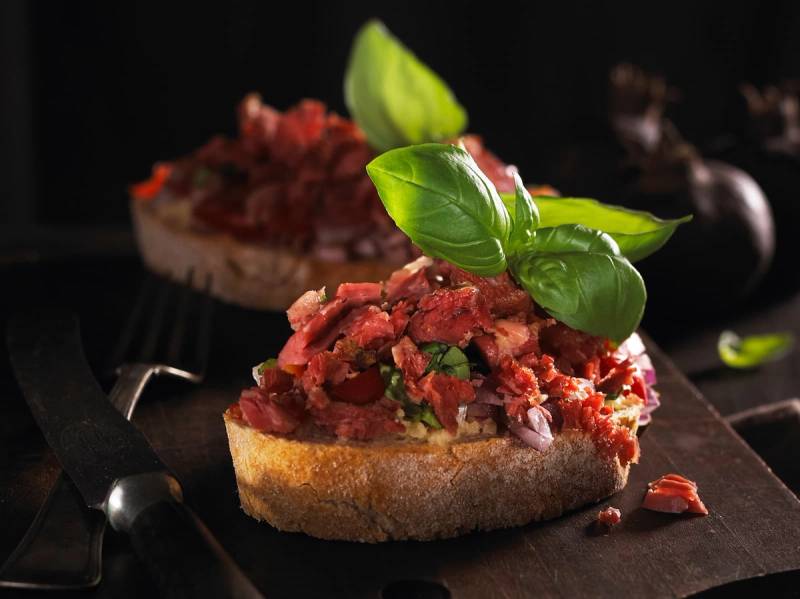 Pinnekjøttbruschetta med hakket pinnekjøtt og friske basilikumblader på en skive brød.