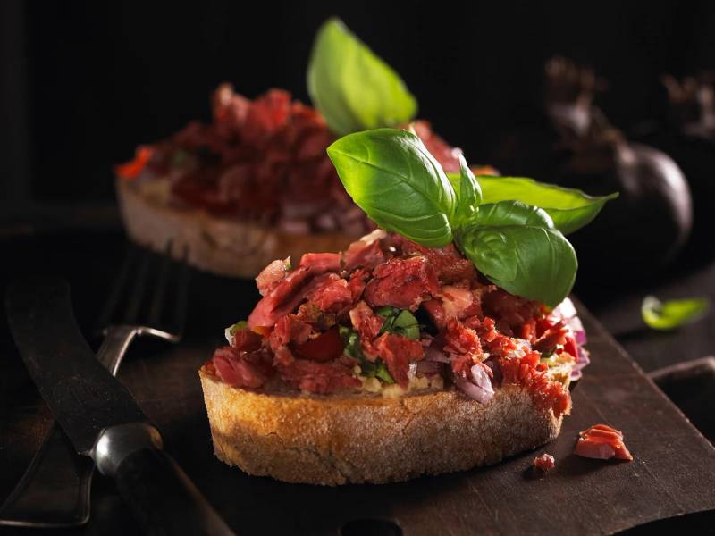 Pinnekjøttbruschetta med hakket pinnekjøtt og friske basilikumblader på en skive brød.