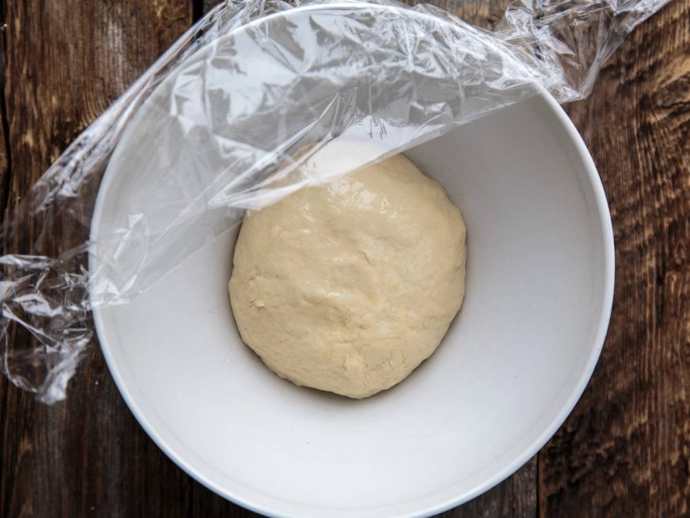 En rund pizzadeig ligger i en hvit bolle med plastfolie over på en trebord.