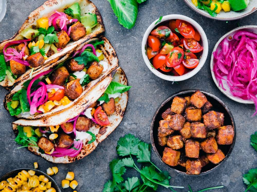 Tofu-tacos med grønnsaker og salsa, omgitt av ulike tilbehør og grønne urter på en stekeplate.