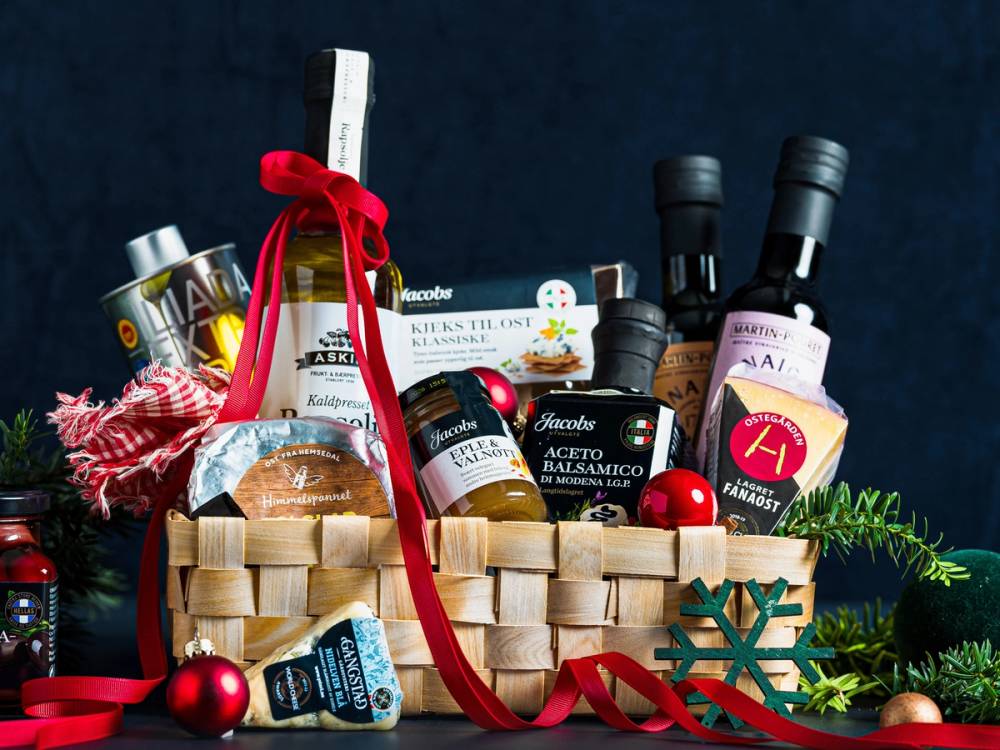 En julegavekurv fylt med gourmetmat og julepynt, omgitt av granbar og julepynt.