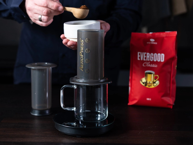 En person lager kaffe med en Aeropress, med en kopp under og en pose med Evergood kaffe ved siden av.