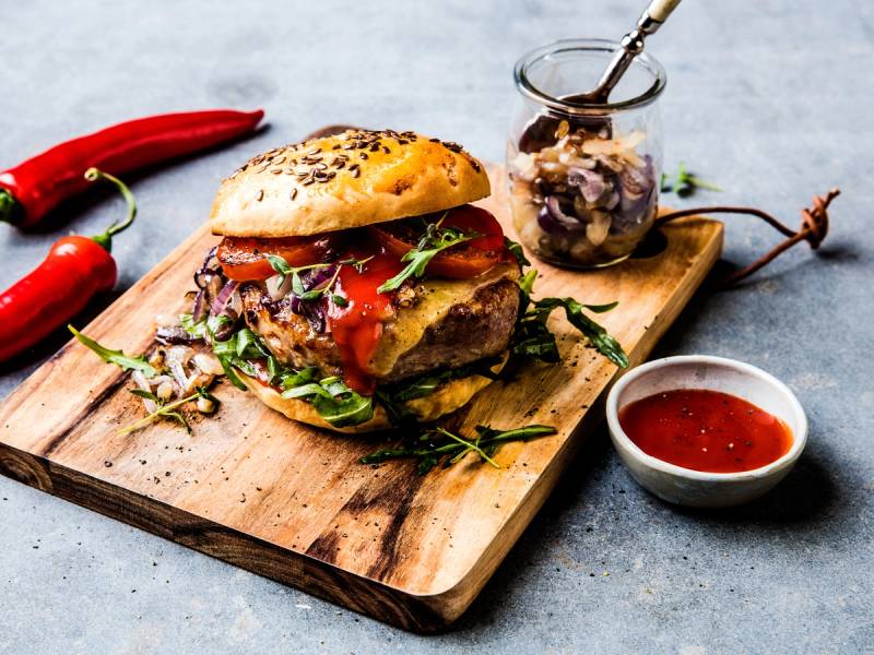 En saftig burger med salat, tomat og kjøtt, servert på en treplate med en liten skål med srirachasaus og en liten glasskrukke med syltetøy eller saus, omgitt av chilipepper på en grå bakgrunn.