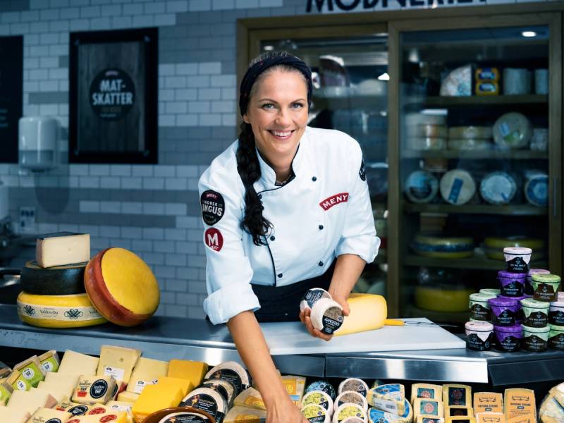 Kvinne i kokkeforkle som selger ost og meieriprodukter i en butikk, med ost og meieriprodukter på disken foran seg.