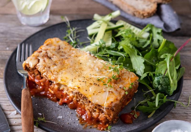 En porsjon lasagne med ost og kjøttsaus på en svart tallerken, servert med en grønn salat ved siden av.