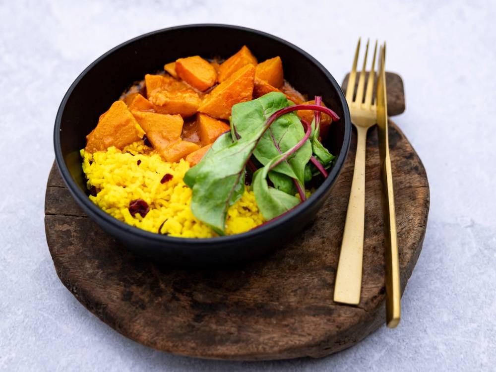 En bol med gult ris, kokte gulerøtter og en liten salat med grønt og rødt, plassert på en rund treplate med gullbestikk ved siden av.