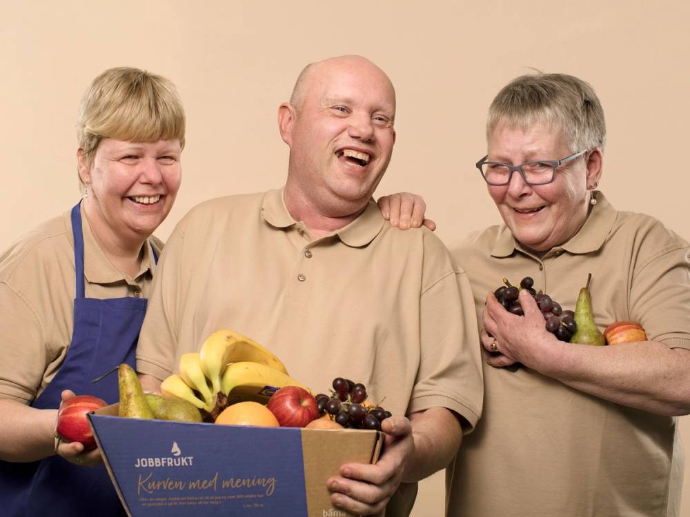 Tre personer med uskarpe ansikter står sammen og holder en eske med frukt, inkludert bananer, epler og druer, i en butikk eller på et kontor.